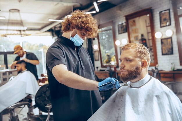 Schließen Sie herauf Mann, der Haarfriseur am Friseursalon trägt Maske während Coronavirus-Pandemie schneidet.