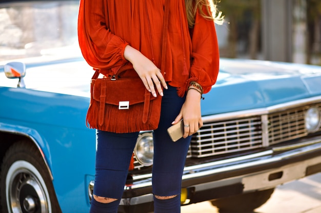 Schließen Sie herauf Modedetails der Frau, die vor Oldtimer, modernem Boho stilvollem trendigem Outfit, Marine-Jeanshose, orange Bluse und Tasche, passenden Accessoires, hält Smartphone, Frühlingssommer aufwirft.