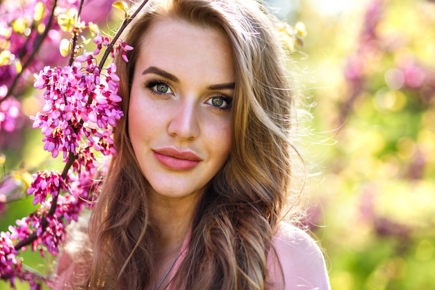 Schließen Sie herauf Modeporträt der zarten eleganten hübschen Frau mit großem grünem Ja und vollen Lippen, natürlichem frischem Make-up und langen flauschigen Haaren, malen ordentlich Sakura blühenden Baum, sonnige Frühlingszeit.