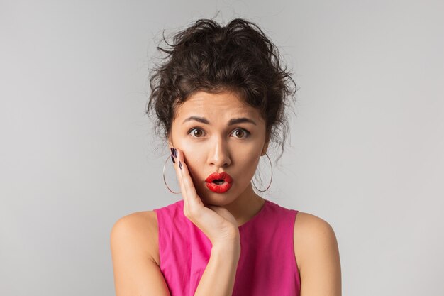 Schließen Sie herauf Porträt der hübschen lustigen Frau mit schockiertem Gesichtsausdruck, junger Hipster, überraschte Emotion, isoliertes, rosa Kleid, Händchenhalten an ihrem Gesicht, große offene Augen, Brünette