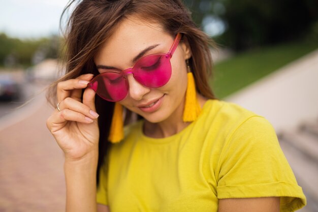 Schließen Sie herauf Porträt der jungen attraktiven lächelnden Frau, die Spaß im Stadtpark hat, positiv, glücklich, tragendes gelbes Oberteil, Ohrringe, rosa Sonnenbrille, Sommerart-Modetrend, stilvolle Accessoires, bunt