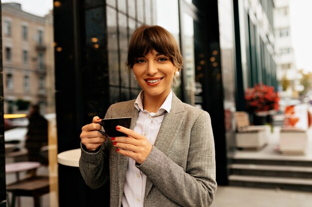 Schließen Sie herauf Porträt der selbstbewussten lächelnden modernen Geschäftsfrau mit einer Tasse Kaffee vor dem Hintergrund eines großen Geschäftsgebäudes Hochwertiges Foto