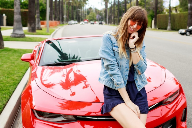 Schließen Sie herauf Porträt der überraschten brünetten Frau, die auf der Motorhaube des erstaunlichen roten Cabrio-Sportwagens in Kalifornien sitzt.