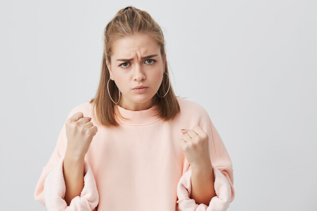 Schließen Sie herauf Porträt der wütenden unzufriedenen europäischen jungen Frau, die ihr Gesicht runzelt, Fäuste vor sich hält, bereit, zu kämpfen oder sich zu verteidigen. Aggressive, emotionale Frau, die sich beleidigt fühlt.