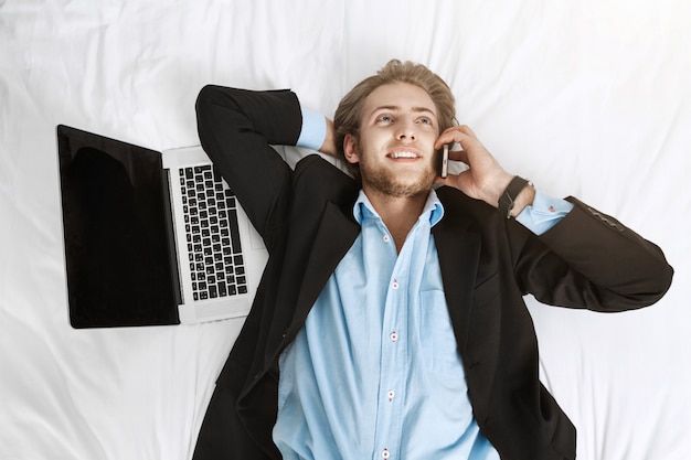 Schließen Sie herauf Porträt des freudigen gutaussehenden Geschäftsmannes, der auf Bett im Anzug mit Laptop und Handy liegt. mit dem Kunden sprechen, sich über seinen Job freuen.