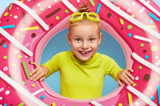 Schließen Sie herauf Porträt des fröhlichen entzückenden Ingwermädchens, das Kopf durch rosa Gummiring steckt, Brille und helles T-Shirt trägt, Sommerferien am Meer verbringt, genießt das Schwimmen. Glückliche Kindheit und Ruhe