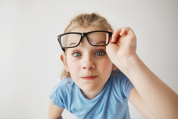 Schließen Sie herauf Porträt des fröhlichen kleinen Mädchens mit blonden Haaren und blauen Augen lustig imitiert erwachsene Person mit Brille mit überraschtem Ausdruck.