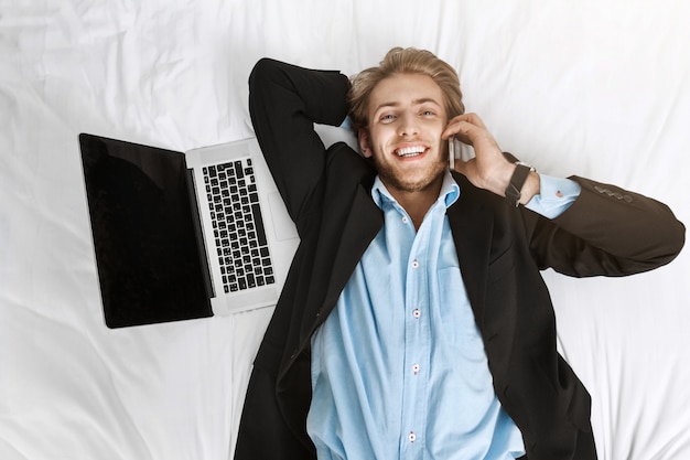 Schließen Sie herauf Porträt des gutaussehenden jungen Geschäftsmannes, der im Bett mit Laptop liegt und am Telefon mit glücklichem Ausdruck spricht.
