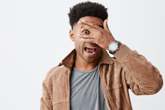 Schließen Sie herauf Porträt des lustigen schönen schwarzhäutigen Mannes mit Afro-Frisur im grauen T-Shirt unter brauner Jacke, die durch Finger in der Kamera mit glücklichem und aufgeregtem Gesichtsausdruck schaut.