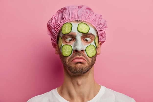 Schließen Sie herauf Porträt des Mannes mit gekreuzten Augen, begierig, perfekte Haut zu haben, trägt pflanzliche Gesichtsmaske und Gurken auf, trägt Duschhaube