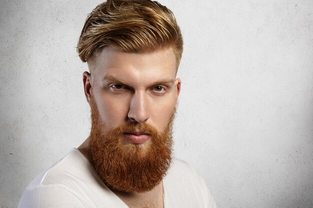 Schließen Sie herauf Porträt des modischen rothaarigen Hipster-Mannes mit unscharfem Bart und trendigem Haarschnitt, der weißes T-Shirt trägt, während lokalisiert gegen Wand mit ernstem oder wütendem Ausdruck aufwirft