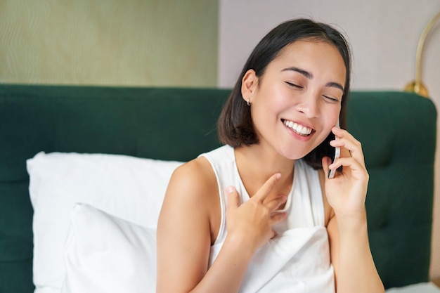 Schließen Sie herauf Porträt des netten asiatischen Mädchens im Bett, das auf Handy mit glücklicher lächelnder Gesichtsfrau wa spricht