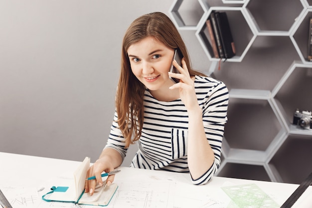 Schließen Sie herauf Porträt des schönen freudigen jungen freien Architekten der schönen sitzenden jungen Tischfrau im Büro, am Telefon mit der Person vom Team sprechend, Arbeitsfehler in Notizbuch aufschreibend