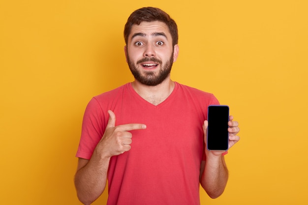 Schließen Sie herauf Porträt des zuversichtlichen gutaussehenden jungen Mannes, der sein Telefon zeigt und mit Zeigefinger auf dem Bildschirm des Geräts zeigt