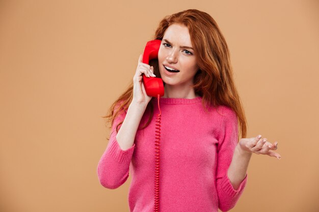 Schließen Sie herauf Porträt eines verwirrten hübschen Rothaarigemädchens, das durch klassisches rotes Telefon spricht