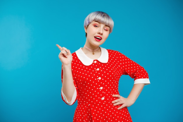 Schließen Sie herauf Porträt schönes dollisches Mädchen mit kurzem hellviolettem Haar, das rotes Kleid über blauer Wand trägt