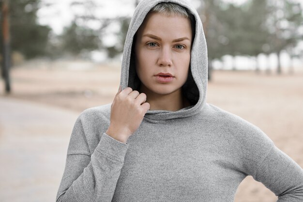 Schließen Sie herauf Schuss von fit selbstbewusster junger Frau, die aktiven gesunden Lebensstil wählt, im Freien trainiert, um perfekte Körperform zu gewinnen und Gewicht zu verlieren, posiert isoliert in stilvollem Hoodie, Blick auf Kamera