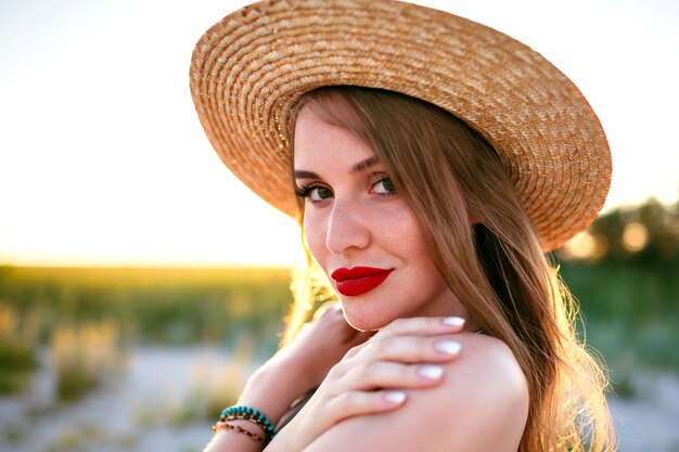 Schließen Sie herauf zartes Porträt der sinnlichen Schönheitsfrau, die auf Feld, Weinlesestil, tragendem Strohhut, Naturschönheitsschminke, sommersprossiges Gesicht und roten vollen Lippen aufwirft.