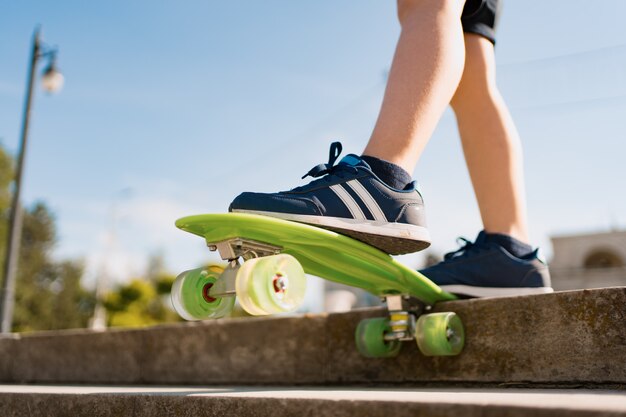 Schließen Sie oben Beine in blauen Turnschuhen, die auf grünem Skateboard in Bewegung reiten. Aktiver urbaner Lebensstil der Jugend, Ausbildung, Hobby, Aktivität. Aktiver Outdoor-Sport für Kinder. Skateboarding für Kinder.