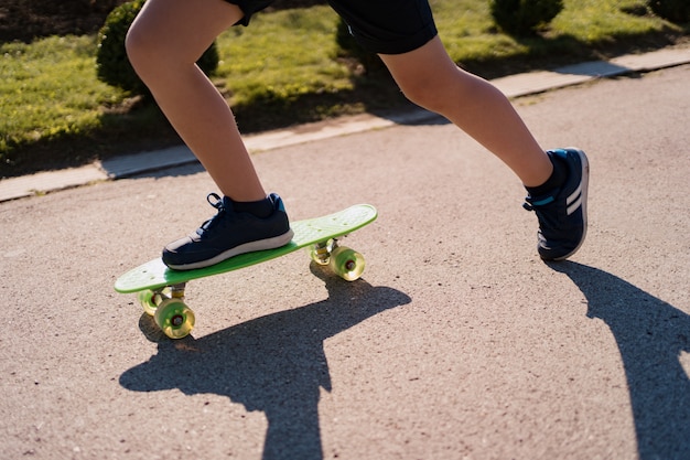 Schließen Sie oben Beine in blauen Turnschuhen, die auf grünem Skateboard in Bewegung reiten. Aktiver urbaner Lebensstil der Jugend, Ausbildung, Hobby, Aktivität. Aktiver Outdoor-Sport für Kinder. Skateboarding für Kinder.