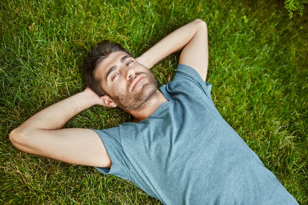 Schließen Sie oben im Freien Porträt des jungen attraktiven reifen bärtigen hispanischen Mannes im blauen T-Shirt, das in der Kamera schaut, auf Boden mit entspanntem Gesichtsausdruck liegend.