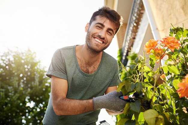 Schließen Sie oben im Freien Porträt des jungen fröhlichen bärtigen Mannes im blauen T-Shirt lächelnd, im Garten mit Werkzeugen arbeitend, Blätter schneidend, Blumen gießend