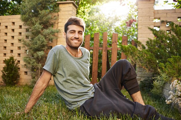 Schließen Sie oben im Freien Porträt des reifen attraktiven bärtigen kaukasischen jungen Mannes im blauen T-Shirt und in der Sporthose lächelnd, auf Gras sitzend, entspannend