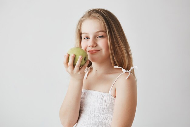 Schließen Sie oben vom lustigen Mädchen mit dem langen blonden Haar, das Apfel in den Händen mit zufriedenem Ausdruck hält, das gesundes Mittagessen in der Schule hava geht.