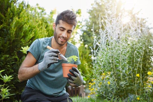 Schließen Sie oben vom reifen bärtigen kaukasischen Mann im blauen T-Shirt, das Blumen in Topf mit Gartenwerkzeugen pflanzt, friedlichen Morgen im Garten verbringen Haus hören.