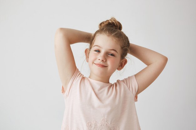 Schließen Sie oben vom süßen kleinen blonden Mädchen mit Brötchenfrisur im rosa lächelnden rosa T-Shirt, Händchenhalten hinter dem Kopf mit glücklichem und zufriedenem Ausdruck.