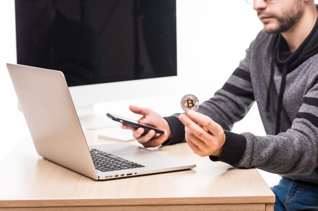 Schließen Sie oben von den Händen des Mannes am Arbeitsplatz mit Laptop und Monitorbildschirm, Telefon und Bitcoin haltend