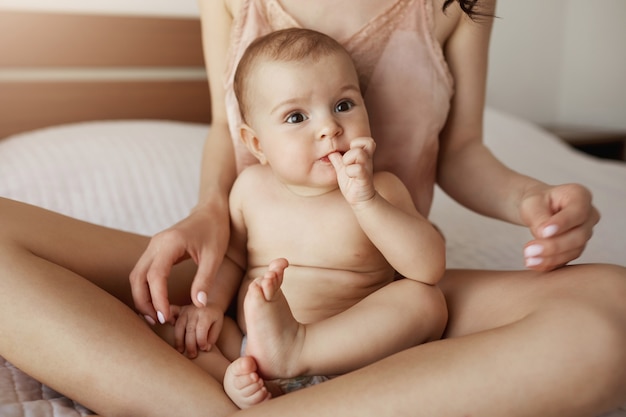 Schließen Sie oben von der jungen Mutter in Nachtwäsche und ihrem neugeborenen Baby, das am Morgen lächelnd auf dem Bett sitzt und zusammen spielt