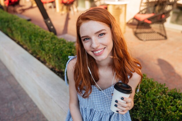 Schließen Sie oben von einem jungen lächelnden rothaarigen Mädchen in den Kopfhörern, die Tasse Kaffee halten