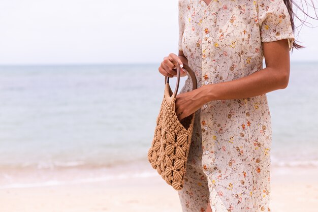 Schließen Sie Schuss der Frau im leichten Sommerfliegerkleid, das gestrickte Tasche am Strand, Meer auf Hintergrund hält.