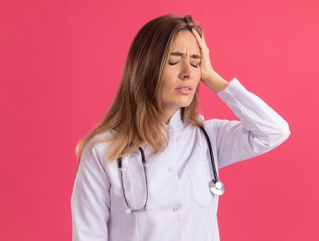 Schmerz mit jungen Augen junge Ärztin, die medizinische Robe mit Stethoskop trägt Hand auf Kopf lokalisiert auf rosa Wand trägt