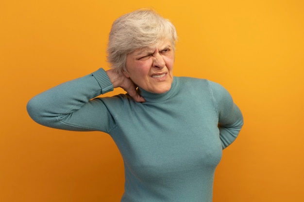 Schmerzende alte Frau mit blauem Rollkragenpullover, die Hand hinter den Hals und auf den Rücken legt und mit einem geschlossenen Auge zur Seite schaut