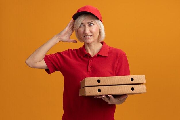 Schmerzende blonde Lieferfrau mittleren Alters in roter Uniform und Mütze, die Pizzapakete hält und auf die Seite schaut, die den Kopf berührt