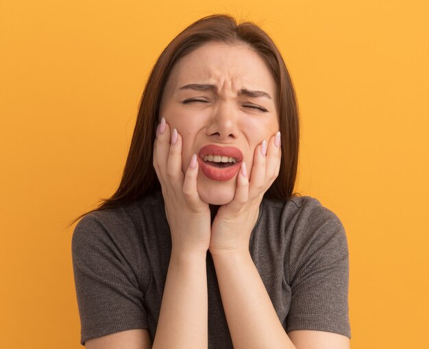 Schmerzende junge hübsche Frau, die mit geschlossenen Augen die Hände auf den Wangen hält und unter Zahnschmerzen leidet