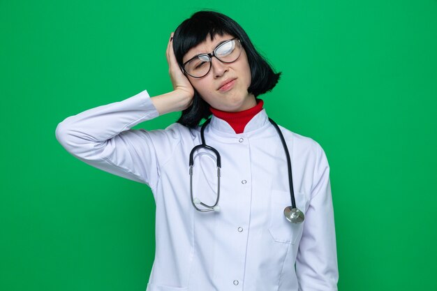 Schmerzende junge hübsche kaukasische Frau mit Brille in Arztuniform mit Stethoskop legt die Hand auf den Kopf