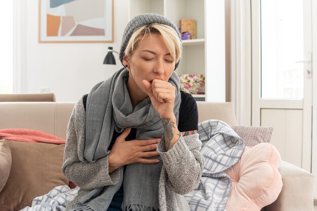 Schmerzende junge kranke slawische Frau mit Schal um den Hals, die Wintermütze trägt und hustet, die Faust nahe am Mund auf der Couch im Wohnzimmer halten