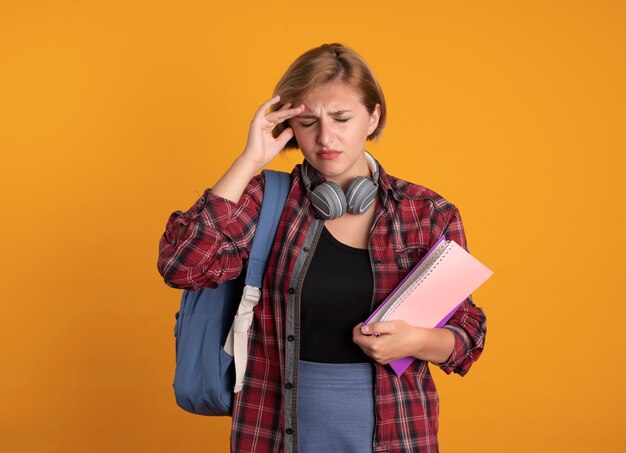 Schmerzende junge slawische Studentin mit Kopfhörern mit Rucksack, die Hand auf die Stirn legt und Buch und Notizbuch hält holding