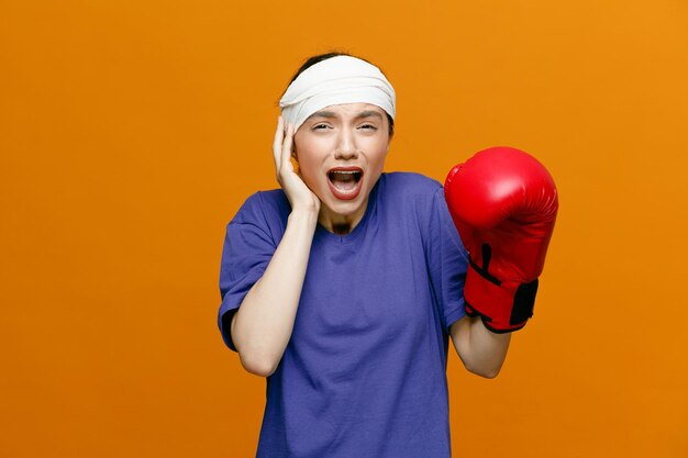 Schmerzende junge sportliche Frau mit T-Shirt und Boxhandschuh, die die Hand auf dem Kopf hält und in die Kamera blickt und eine andere Hand in der Luft hält, wobei der Kopf mit einem Verband isoliert auf orangefarbenem Hintergrund umwickelt ist