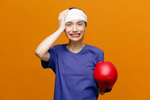 Schmerzende junge sportliche Frau mit T-Shirt und Boxhandschuh, die in die Kamera blickt und die Hand auf dem Kopf und eine andere Hand in der Luft hält, wobei der Kopf mit einem Verband isoliert auf orangefarbenem Hintergrund umwickelt ist