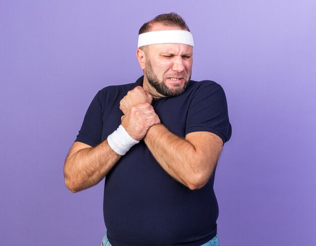 schmerzender erwachsener slawischer sportlicher Mann mit Stirnband und Armbändern, der die Hand isoliert auf lila Wand mit Kopienraum hält