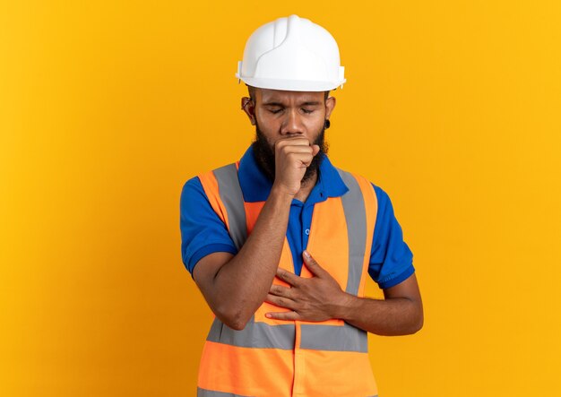 schmerzender junger Baumeister in Uniform mit Schutzhelm, der isoliert auf orangefarbener Wand mit Kopienraum hustet