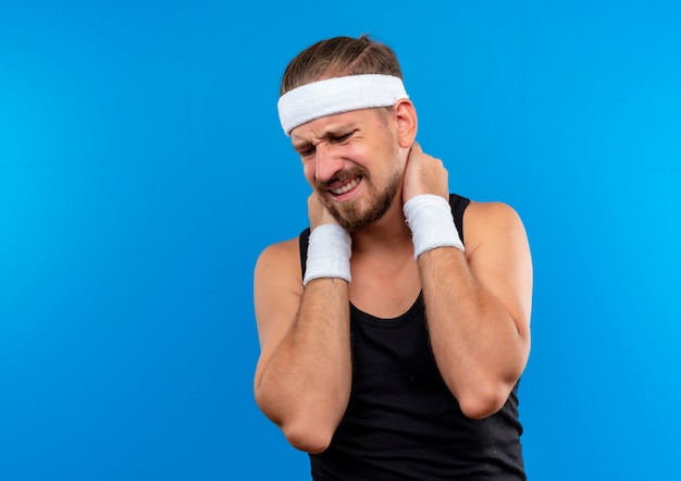 Schmerzender junger, gutaussehender, sportlicher Mann mit Stirnband und Armbändern, der die Hände auf den Hals legt und isoliert auf blaue Wand mit Kopierraum schaut