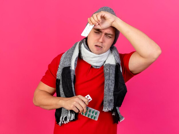 Schmerzender junger hübscher blonder kranker Mann, der Wintermütze und Schal hält Packungen der medizinischen Pillen, die unten schauen und Kopf lokalisiert auf rosa Wand halten