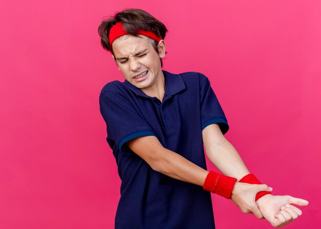 Schmerzender junger hübscher sportlicher Junge, der Stirnband und Armbänder mit Zahnspangen trägt, die Handgelenk lokalisiert auf purpurroter Wand mit Kopienraum halten