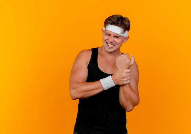 Schmerzender junger hübscher sportlicher Mann, der Stirnband und Armbänder hält, hält Handgelenk lokalisiert auf Orange mit Kopienraum
