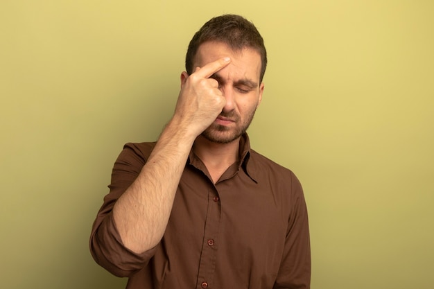 Schmerzender junger kaukasischer Mann, der Finger auf Stirn mit geschlossenen Augen zeigt, die Kopfschmerzen lokalisiert auf olivgrünem Hintergrund mit Kopienraum haben